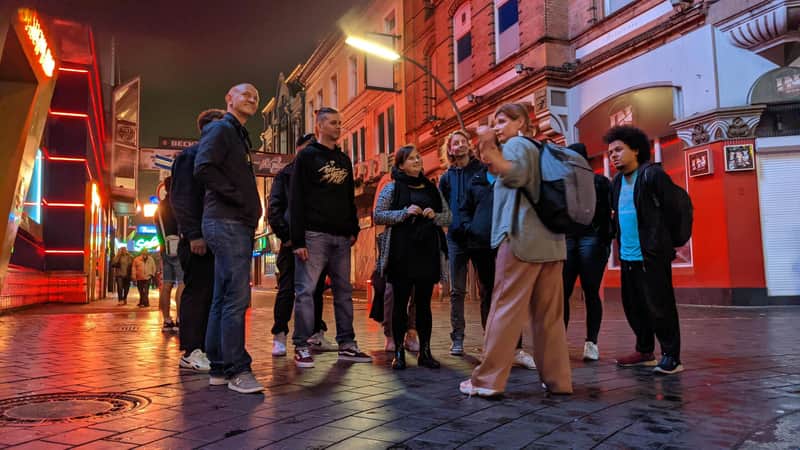 A woman guiding a group of people on a city street near red-lit buildings at dusk.