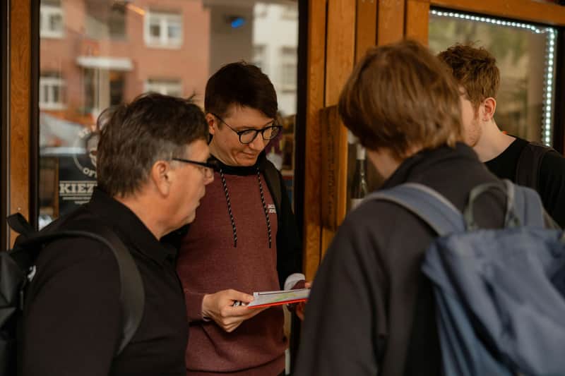 Three people stand outside a shop; one man reads a pamphlet while the others look on.