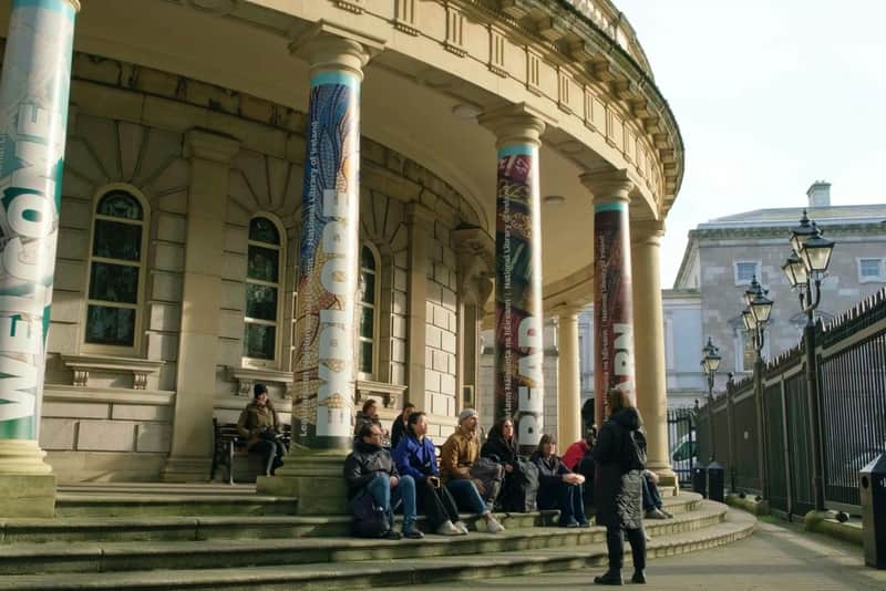 Dublin: Guided Dark Walking Tour (Dublin) People sitting and talking on the steps of a building with large columns featuring the words "EXPLORE," "READ," and "LEARN."