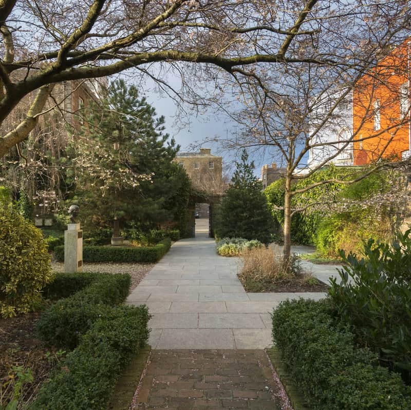 Early Access: Book of Kells & Dublin Castle Tour (Dublin) A garden pathway lined with hedges and trees, leading to a building in the background. Bust sculpture is visible on the left.