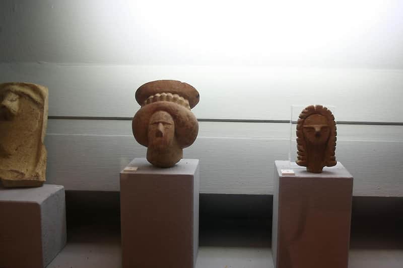 Petite sculpture en pierre d'une tête avec une coiffe, présentée sur un piédestal, sous un éclairage ciblé dans un musée.
