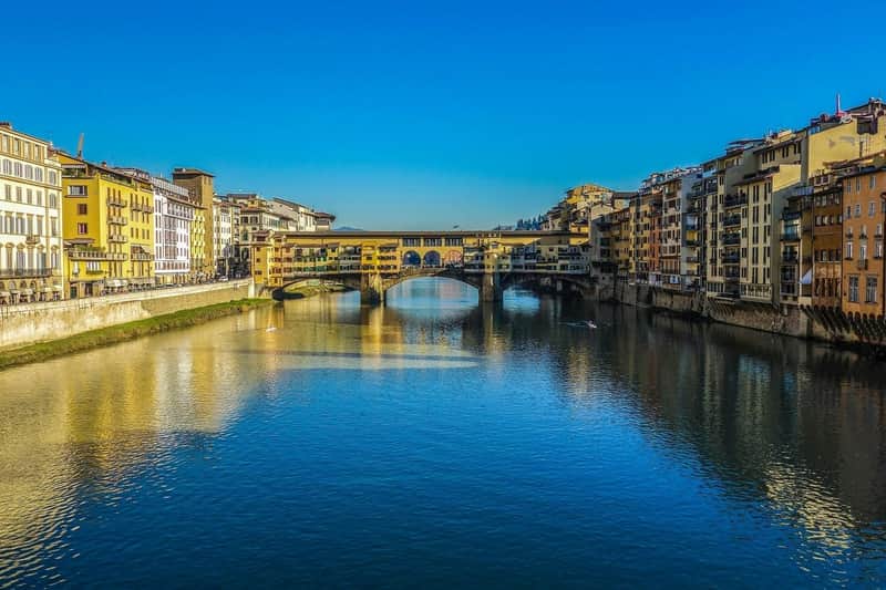 Un pont historique enjambant une rivière paisible, bordé de bâtiments de part et d'autre, sous un ciel bleu limpide.