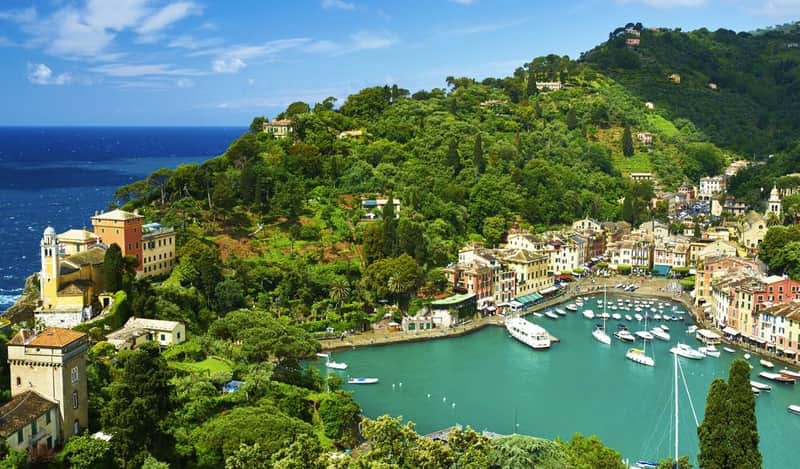 A coastal town with colorful buildings, yachts docked in a turquoise harbor, surrounded by lush green hills and blue sky.