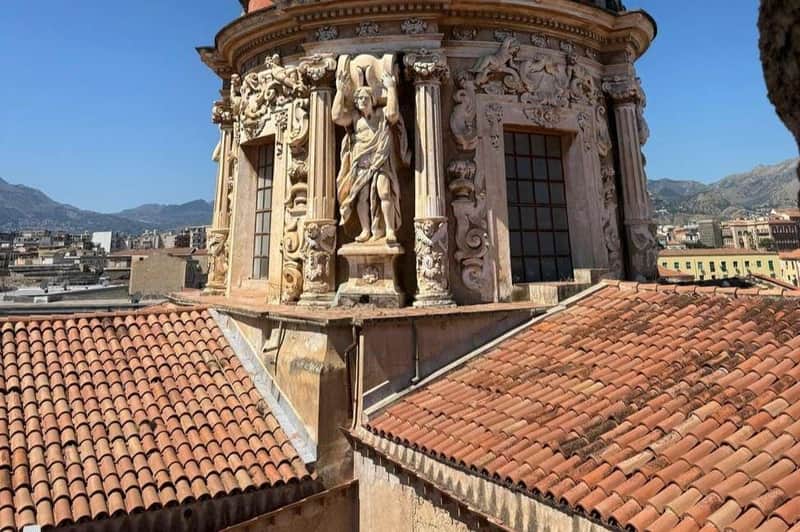 Ornate stone building with statues and columns, alongside red-tiled rooftops. The background includes distant mountains and sky.
