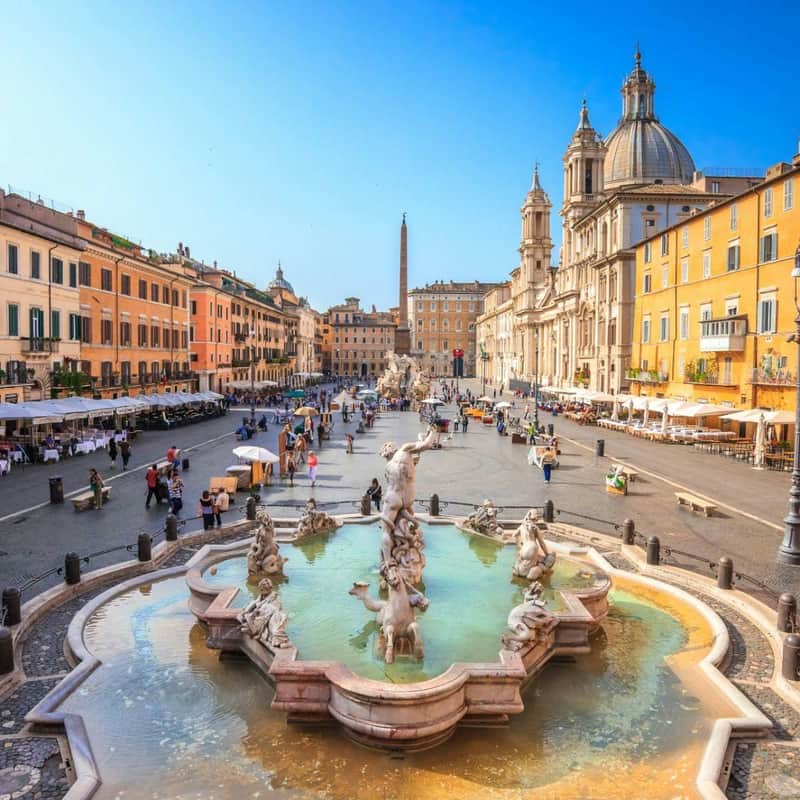 A lively square with a central fountain, surrounded by historic buildings, cafes, and people walking and sitting.