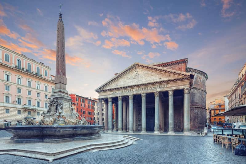 Ancient Roman Pantheon with columns and triangular pediment, adjacent to a fountain and obelisk, under a twilight sky.
