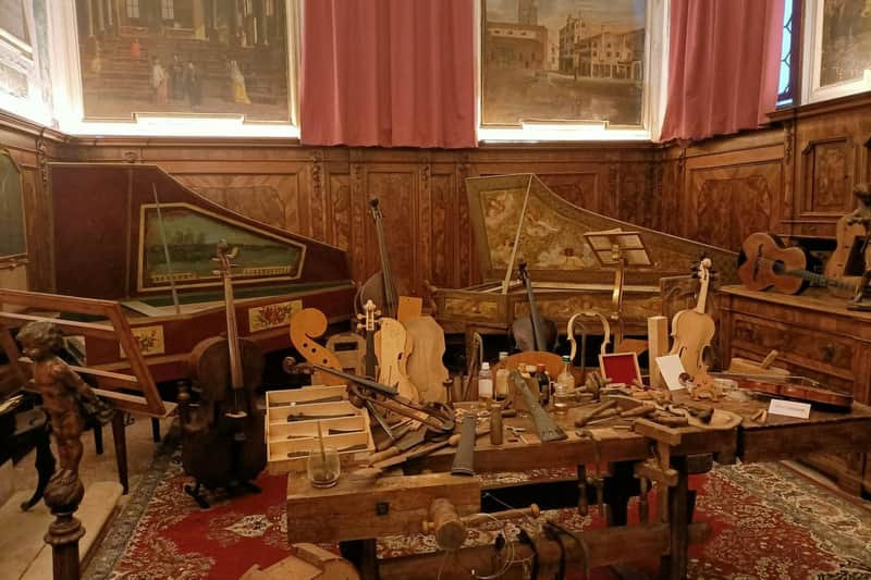 A workshop filled with string instruments, including violins and harpsichords, surrounded by various woodworking tools and parts.