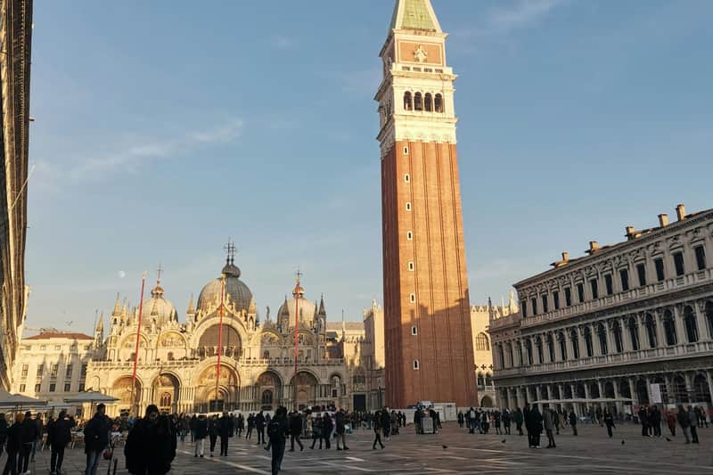 A tall clock tower and ornate cathedral fronting a busy public square under a clear blue sky, with people walking around.
