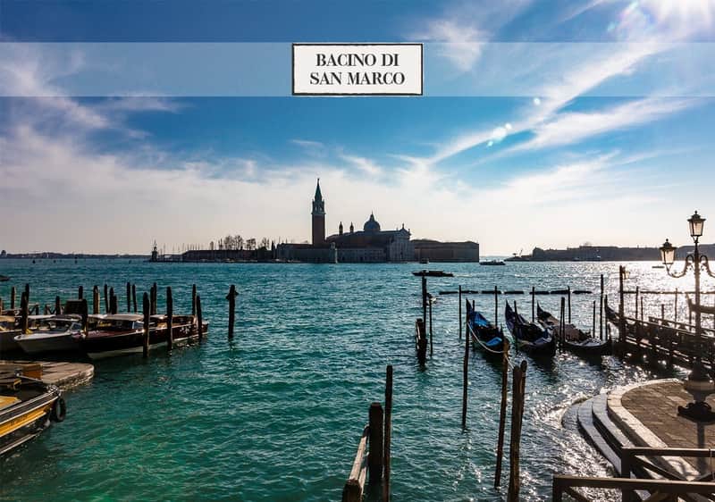 Venice: St. Mark's Square Historical City Walking Tour Waterfront view with blue water, docked gondolas, and a distant island with a tall bell tower and historic buildings beneath a bright sky.