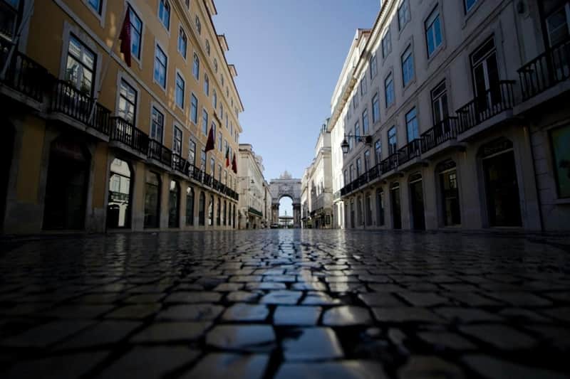 Low angle view of a cobblestone street flanked by buildings with balconies, leading to an archway under a clear blue sky.