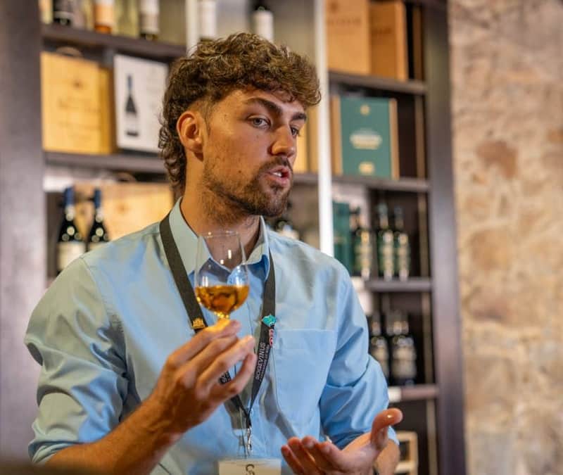 Porto City Tour Half-Day: Guided Visit with Wine Cellar and Tastings (Porto) A person with a blue shirt and lanyard holds a glass of wine, speaking in a room lined with wine bottles and boxes.