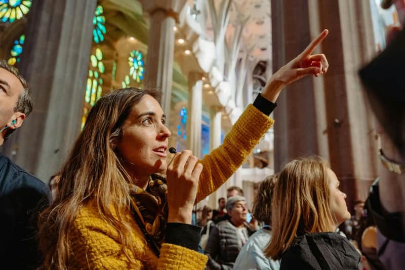 Eine Frau in einer gelben Jacke spricht in ein Mikrofon, während sie in einer Kathedrale mit Buntglasfenstern nach oben zeigt.