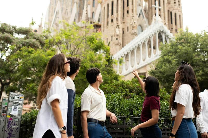 Hay tres personas de pie en un parque, mirando hacia arriba, con la Sagrada Familia al fondo, rodeadas de vegetación y elementos urbanos.
