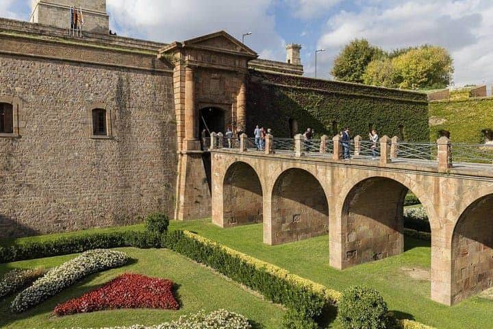 Steinbrücke, die zu einem alten Festungseingang führt, auf der Menschen spazieren gehen. Manicured Gärten mit bunten Blumen sind unten.
