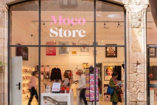 ガラス張りのファサードに、アートやグッズが飾られた「Moco Store」。 人が外を歩いている。
