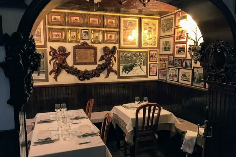 マドリード:プラド美術館+ウォーキングツアー+「Sobrino de Botín」でのランチ 2つのセットテーブルの上に天使の彫刻が飾られた、壁に額装された絵とアートワークが飾られた居心地の良いレストランのコーナー。