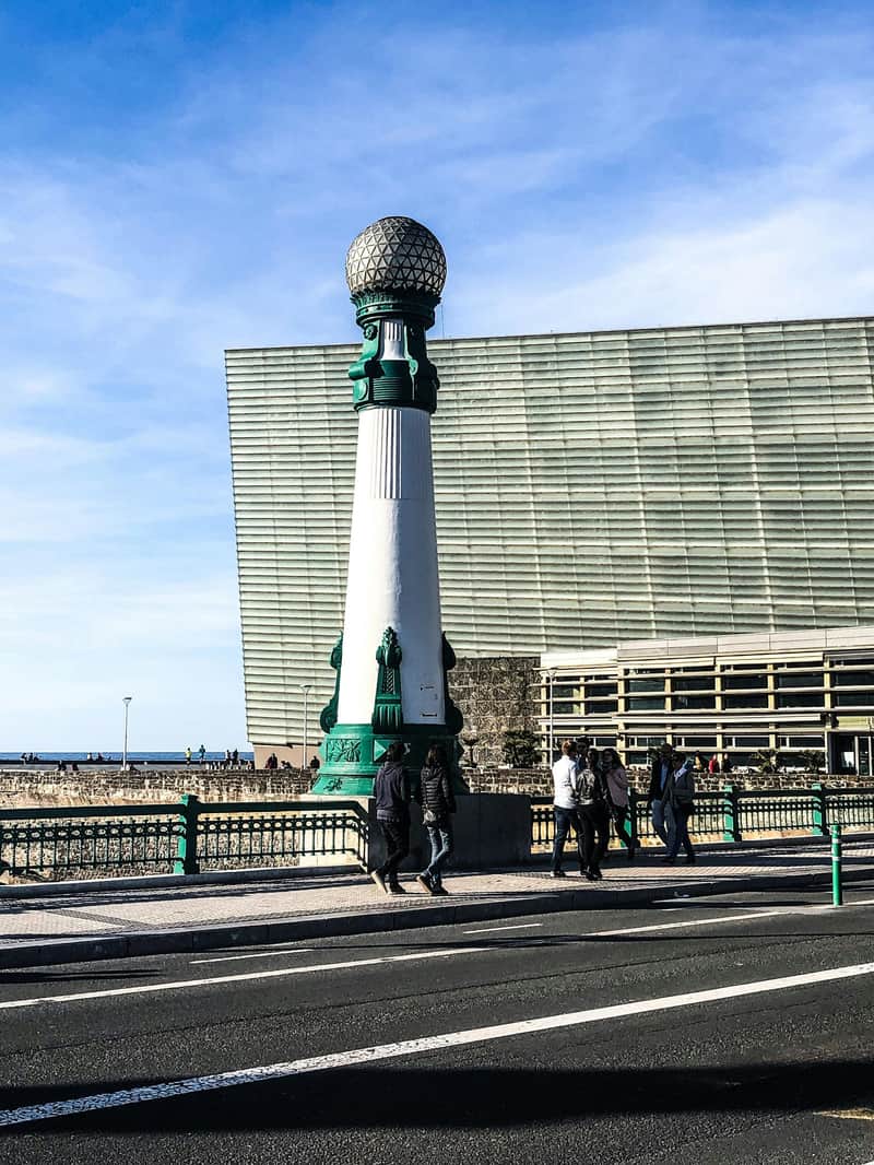 San Sebastián Historic & Cultural Walking Tour (San Sebastián) People walk by a tall, ornate green and white streetlight on a bridge, with a large modern building and the ocean in the background.