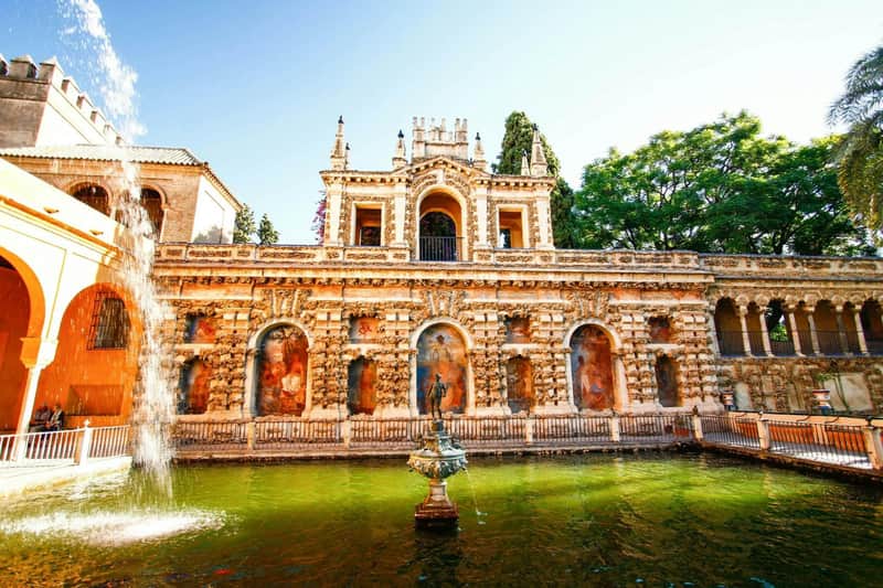 Ornate historic building with arches and statues reflected in a green pond, surrounded by trees under a clear sky.