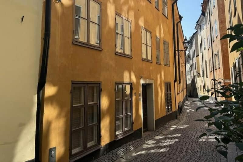 Stockholm: ABBA Small Group Guided Walking Tour A narrow cobblestone street lined with yellow plastered buildings featuring multiple windows and a shadow pattern on the walls.