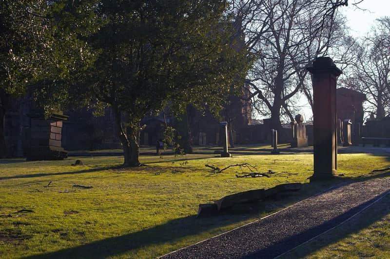 A sunlit graveyard with trees, gravestones, and a pathway, showcasing a calm and serene atmosphere.