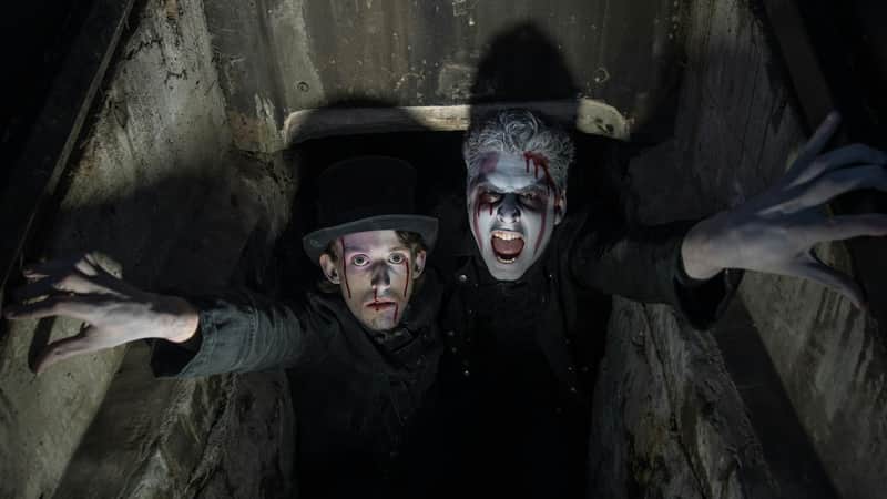 Two people with dramatic makeup and fake blood look upwards from a dark, enclosed space; one wears a top hat.