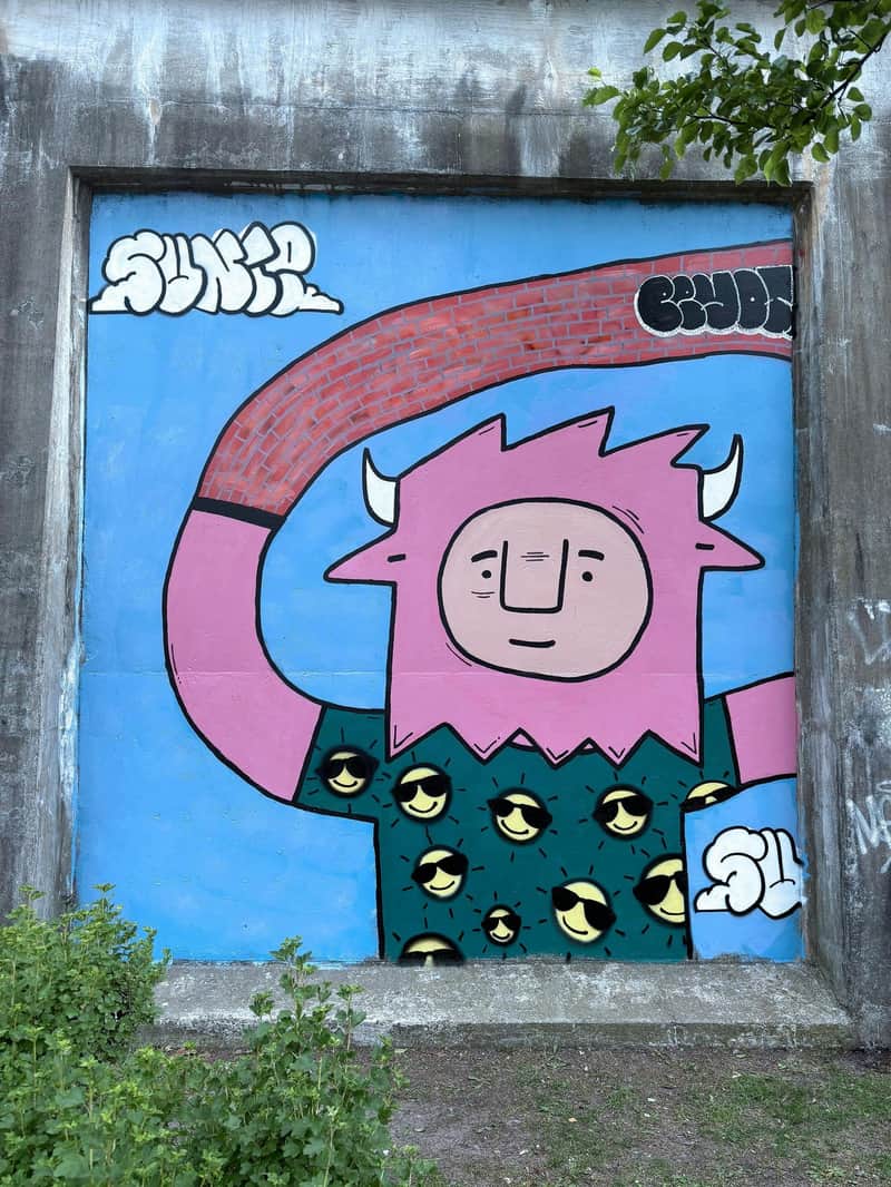 Graffiti coloré représentant un personnage de dessin animé rose avec des cornes, portant un t-shirt orné de smileys jaunes, sur fond bleu.