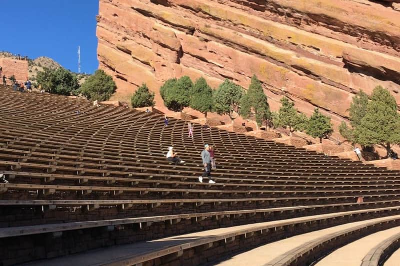 Denver : Red Rocks Park & Amphitheatre - Visite guidée en petit groupe Des gens marchent et s'assoient sur des gradins en pierre, avec en toile de fond de grandes formations rocheuses rouges et des arbres clairsemés, sous un ciel bleu clair.