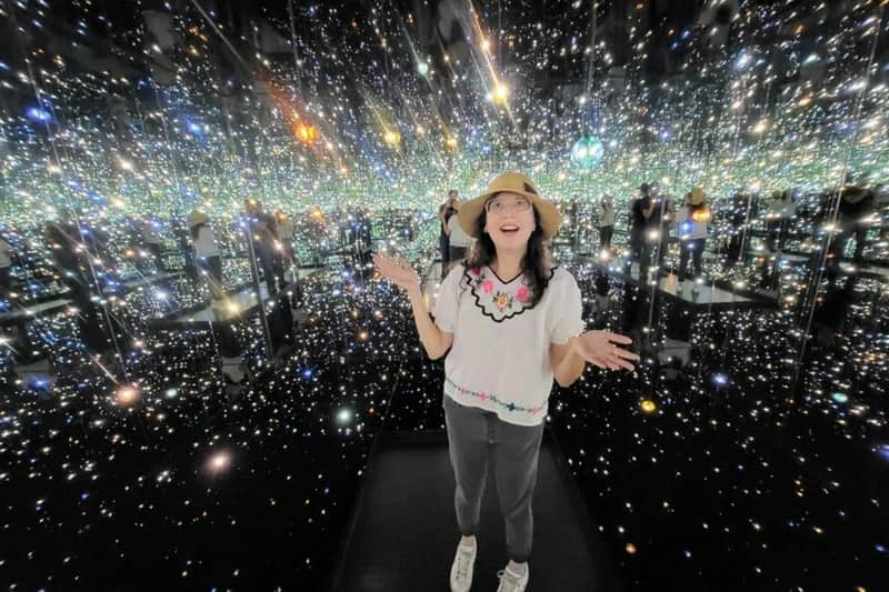 Los Angeles: Downtown Art & Music Guided Walking Tour (Los Angeles) A woman stands in a room filled with mirrored walls and twinkling lights, wearing a hat, glasses, floral top, gray pants, and sneakers.