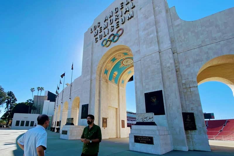 Los Angeles: Exposition Park Guided Walking Tour (Los Angeles) Two people stand outside the Los Angeles Memorial Coliseum, featuring Olympic rings and large archways, on a clear day.