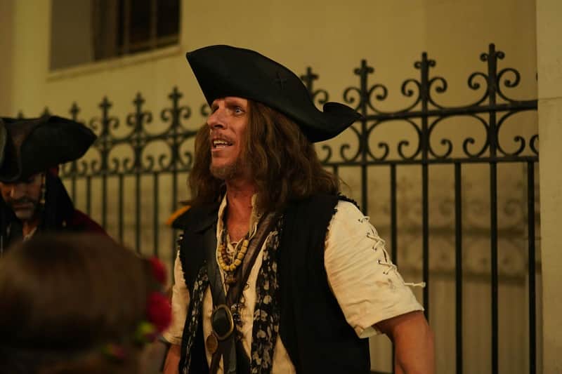Una persona vestita da pirata con un cappello a tricorno e i capelli lunghi, in piedi davanti a una recinzione decorativa in metallo.