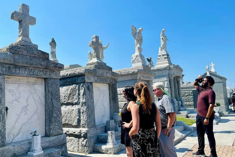New Orleans: Guided Bus Tour + Garden District Walk Three people standing in front of ornate stone tombs with statues in a sunny cemetery.