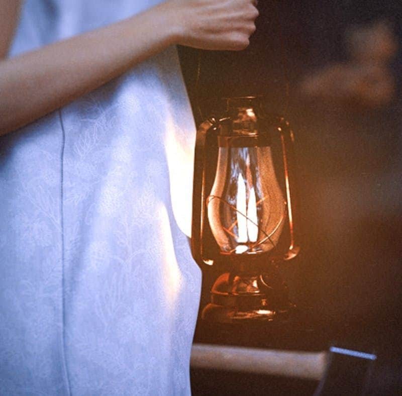 The Dim Corner Tour Person in a light blue dress holding a glowing lantern in a dimly lit setting.
