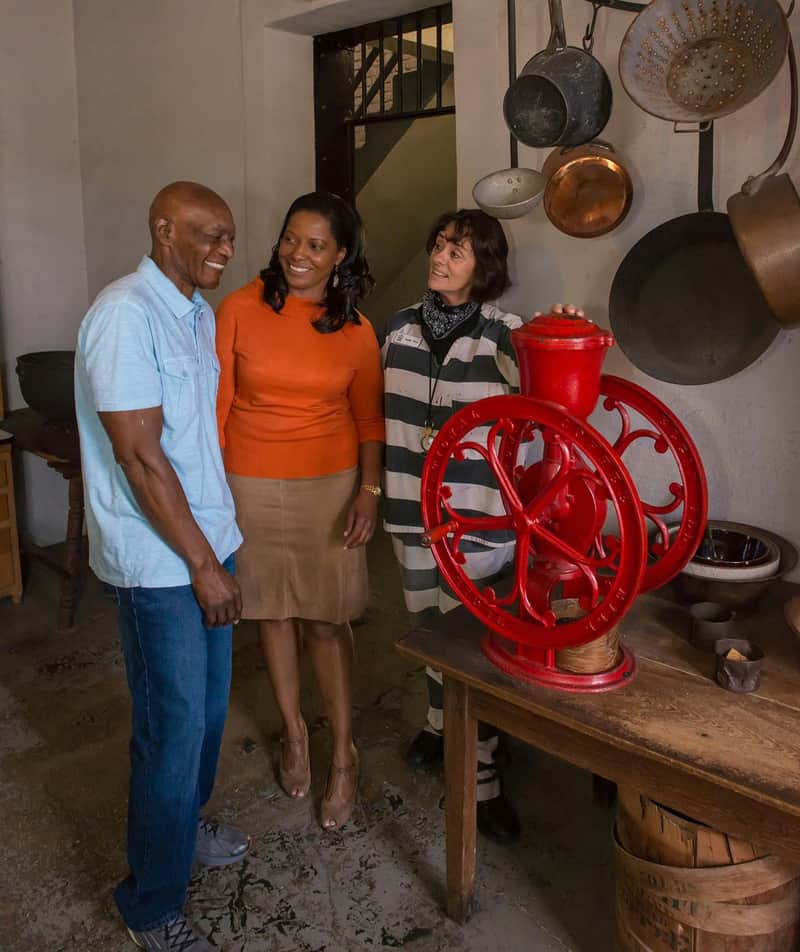 Three people stand inside a rustic kitchen, conversing near a vintage red coffee grinder and hanging pots.