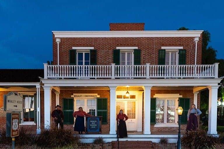 Whaley House Museum: Haunted Guided Evening Tour A historic brick building with a white balcony and green shutters, featuring three costumed figures on the porch at night.